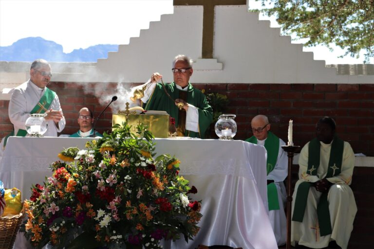 Elderly priest in eyewear and catholic vestment standing with censer near table with Bible and diverse deacons during mass process near colorful bouquet in sunlight in back lit